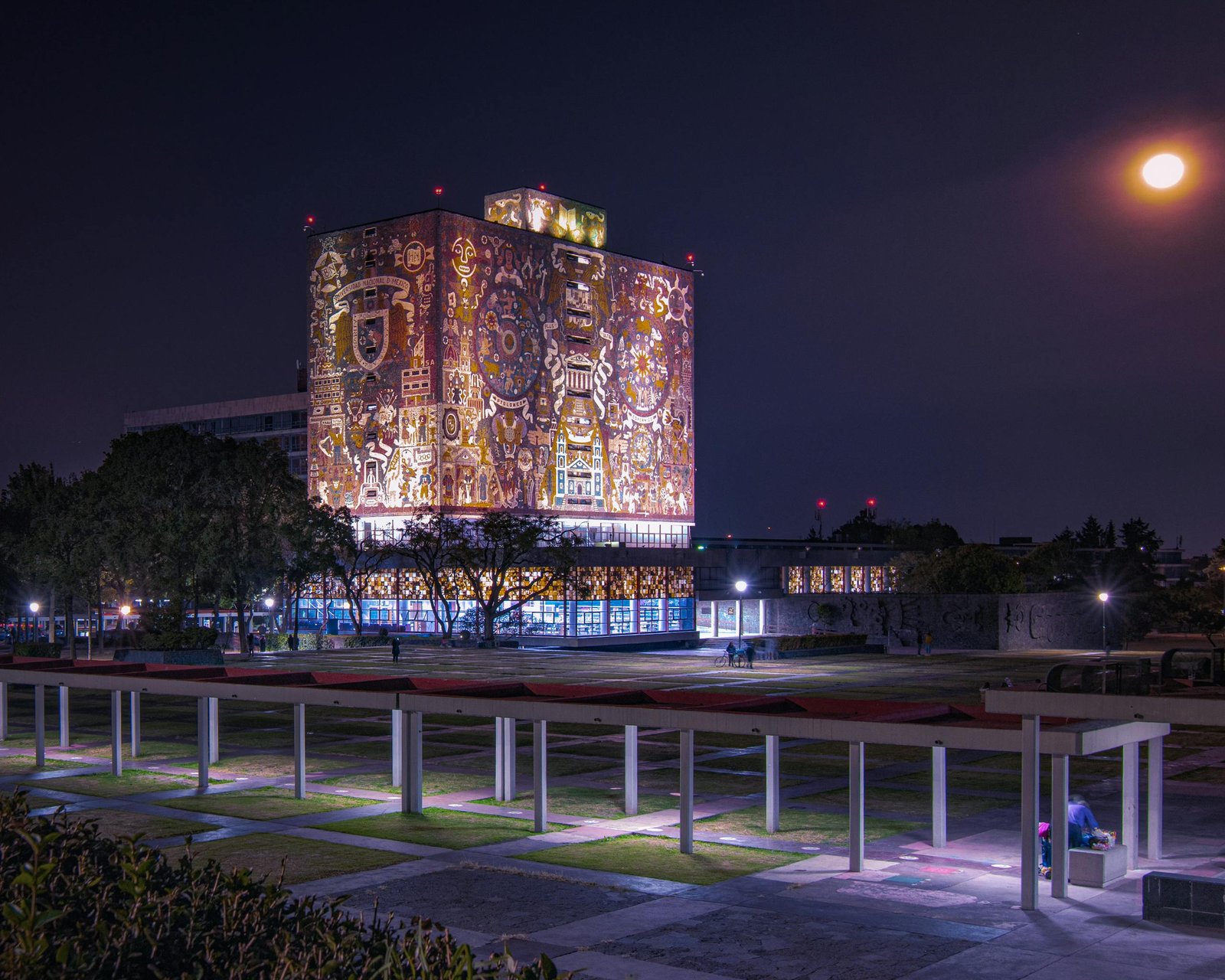 Illuminated UNAM Central Library with vibrant murals and full moon in Mexico City.